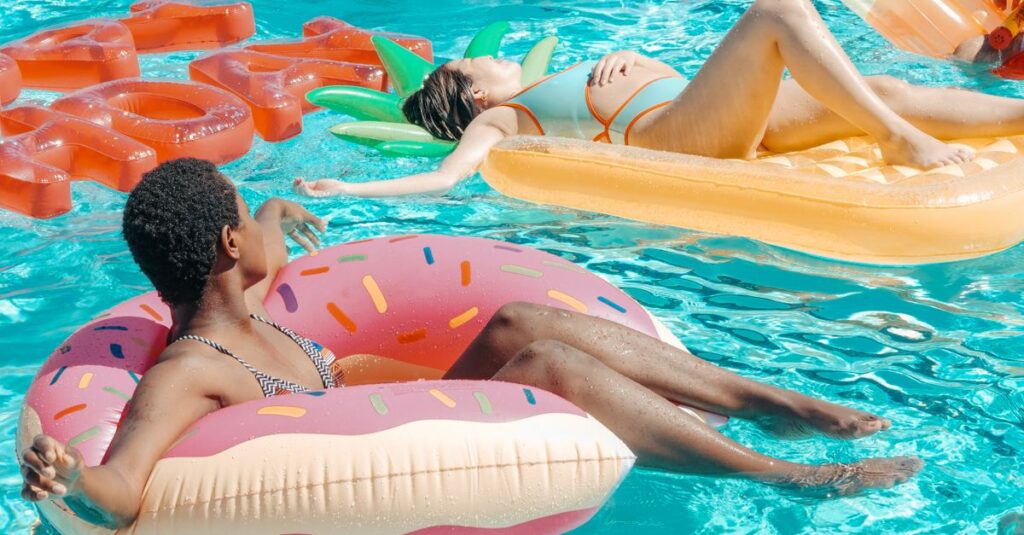 Duas mulheres em boias de piscina, uma em forma de donut e outra em formato de abacaxi, relaxando na água.